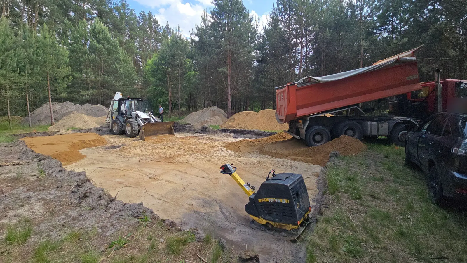 Wywrotka dowozi kruszywo, na podbudowie zagęszczarka płytowa, w tle koparko-ładowarka i las