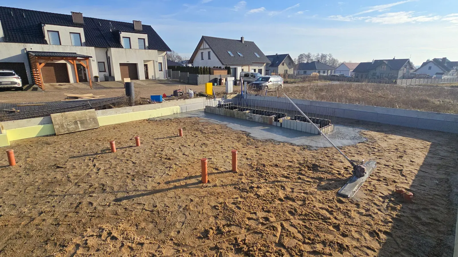 Kształtki brzegowe na obwodzie płyty i piony instalacji wyprowadzone z piaskowej podbudowy, w tle domy
