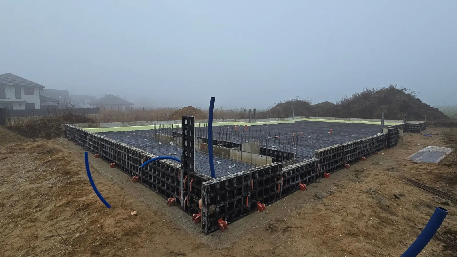 Płyta fundamentowa w szalunku systemowym z wyprowadzonymi niebieskimi przepustami instalacyjnymi, mglisty dzień