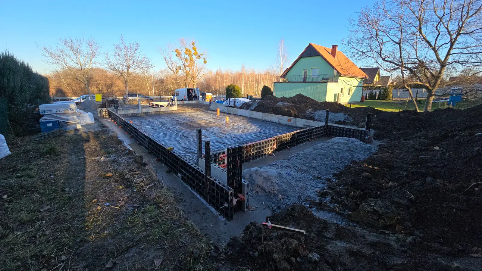 Płyta fundamentowa w szalunku systemowym przed betonowaniem, w tle dom jednorodzinny